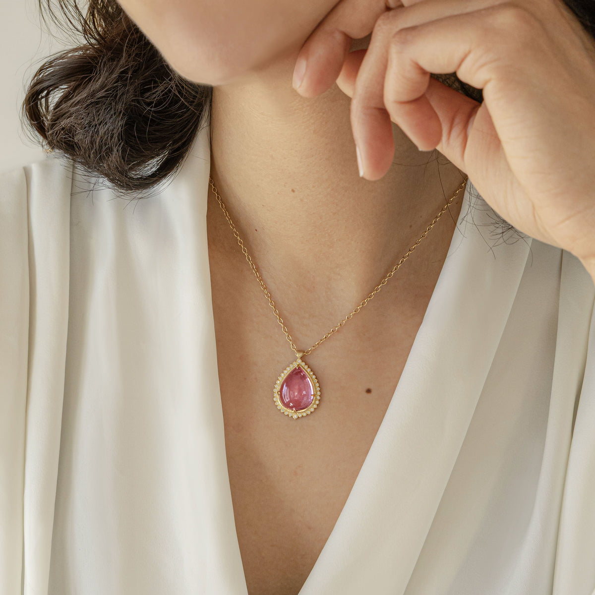 A close-up lifestyle shot of a woman wearing a gold chain necklace with a teardrop-shaped pink gemstone pendant, styled with a white satin blouse.